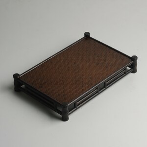 Puede incluir: Una bandeja rectangular de madera con una parte superior de bamb&uacute; tejido. La bandeja tiene un acabado marr&oacute;n oscuro y se eleva sobre cuatro patas.