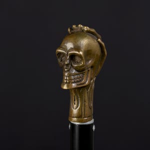 Peut inclure: Une poignée de canne en métal couleur bronze en forme de crâne humain avec une main posée sur le sommet du crâne. Le crâne présente des orbites creuses, une cavité nasale et une rangée de dents visible. Il est fixé sur un manche cylindrique noir.