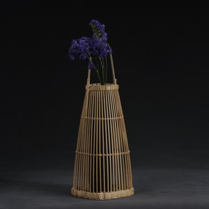 May include: A tall, cylindrical bamboo vase with a natural finish. The vase has a woven design and is filled with purple flowers.