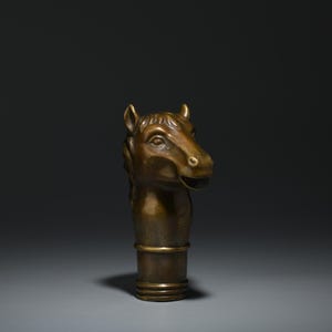 May include: A bronze-colored horse head sculpture. The horse head is detailed with visible eyes, ears, and a slightly open mouth. The sculpture has a cylindrical base with a gold-colored band. The background is a gradient of dark gray.