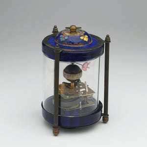 Puede incluir: Un reloj azul y dorado con una cúpula de vidrio y un globo giratorio en su interior. La esfera del reloj es visible a través del globo y tiene números romanos. La cúpula está decorada con flores de esmalte de colores.