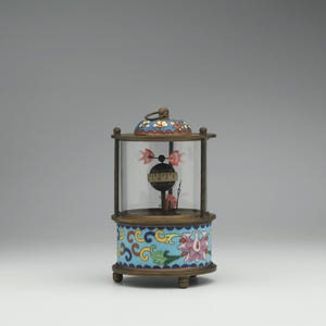 May include: A small, round, cloisonné clock with a glass dome. The clock face is visible through the dome and has black Roman numerals. The clock is decorated with colorful floral patterns and is sitting on a white surface.
