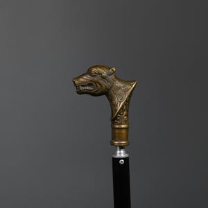 May include: A walking cane with a decorative handle. The handle is shaped like a snarling bear's head, cast in a bronze-colored metal. The shaft is black, and the cane has a brass-colored collar.