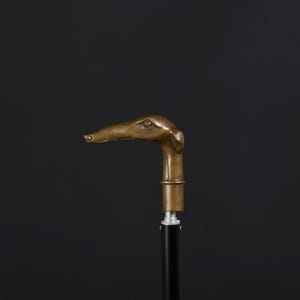 May include: A walking stick featuring a bronze-colored handle shaped like a greyhound dog head. The handle is attached to a smooth, black cylindrical shaft with a silver-toned metal collar connecting the two distinct sections of the cane.