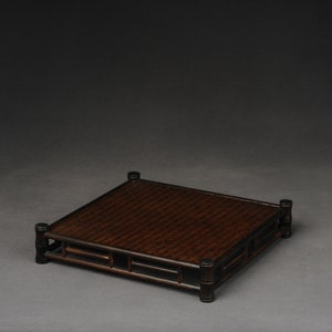 Op de afbeelding: Een vierkante, donkerbruine bamboestandaard met een geweven bovenkant. De standaard heeft een eenvoudig, geometrisch ontwerp met vier poten en een rechthoekig frame.