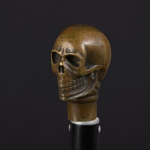 Könnte beinhalten: Eine bronzefarbene Metallschädel-Skulptur als Griffaufsatz mit detaillierten Augenhöhlen, Nasenhöhle und ausgeprägter Kieferpartie. Der Schädel ist auf einem glatten, zylindrischen schwarzen Schaft montiert, der mit zwei sichtbaren silbernen Schrauben befestigt ist.
