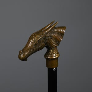 May include: Bronze dragon head sculpture with intricate scale and armor detailing. The dragon head is mounted on a black pole. The sculpture is a decorative item, possibly a cane or staff topper.