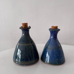 May include: Two ceramic bottles with cork stoppers. The bottles are a deep blue with a glossy finish and a bulbous shape. The corks are a light brown colour. The bottles are likely for oil or vinegar.