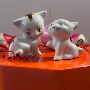 Può includere: Due statuette di gatto in ceramica bianca con occhi gialli e segni neri. Un gatto ha una macchia nera e oro sulla testa. I gatti sono su una superficie arancione con nastro rosso e ornamenti rosa sullo sfondo.
