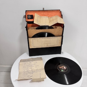 Peut inclure: Un porte-disques vinyle noir vintage avec un couvercle ouvert, révélant une doublure orange et des séparateurs de disques. Une feuille de papier manuscrite et un disque vinyle noir sont sur une table blanche. Le porte-disques est étiqueté "The Lifeguard Carrier".