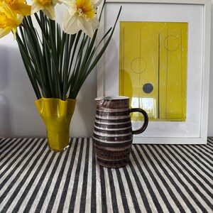 May include: A brown and white striped ceramic jug with a handle. The jug is sitting on a black and white striped tablecloth. A bouquet of yellow daffodils is in a yellow vase in the background.