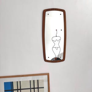 Peut inclure: Un miroir rectangulaire avec un cadre en bois. Le miroir présente un contour décoratif en fil de fer d'une forme féminine. Le cadre est de couleur marron moyen. Le miroir est fixé sur un mur blanc.