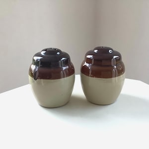 May include: A pair of vintage salt and pepper shakers. Each shaker is a small, rounded jar with a dark brown top, a medium brown band, and a cream-colored base. The tops have small holes for dispensing the contents.