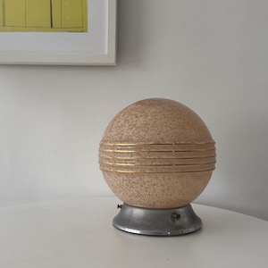 May include: A vintage, spherical table lamp with a speckled, peach-colored glass shade. The shade is accented with gold bands and sits atop a silver-toned metal base. The lamp is a decorative lighting fixture.