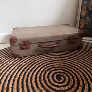Pode incluir: Uma mala vintage com um exterior bege texturizado e detalhes em couro castanho. A mala tem uma pega e fechos metálicos. Os cantos são reforçados com couro castanho. A mala está sobre um tapete redondo com um padrão espiral preto e bege.