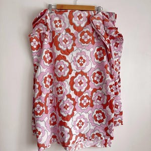 Op de afbeelding: Een bloemmotiefstof in rood, roze en wit, hangend aan een houten hanger. De stof heeft een herhalend bloemdesign met een retro-esthetiek. De stof is waarschijnlijk een textiel voor kleding of woondecoratie.