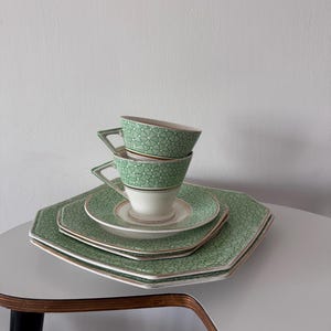 Könnte beinhalten: Ein grünes und weißes Teeservice mit geometrischem Muster, bestehend aus Tassen, Untertassen und Tellern. Das Set hat einen goldenen Rand. Die Tassen sind gestapelt. Das Geschirr ist auf einem weißen Tisch arrangiert.