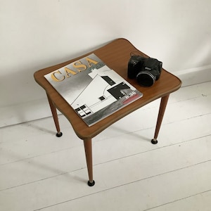 Op de afbeelding: Een moderne bijzettafel uit het midden van de eeuw met een houten blad en vier poten. De tafel heeft een licht gebogen blad en is gemaakt van licht hout. Op de tafel ligt een tijdschrift met de titel "Casa" en een zwarte camera.