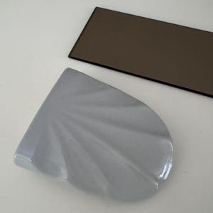 May include: A light gray ceramic object with a fan-shaped, scalloped design featuring raised ridges radiating from a straight edge. Beside it lies a thin, rectangular, dark brown glass piece with a smooth, reflective surface and dark edges.