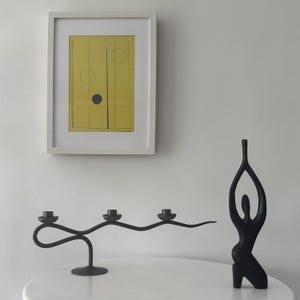 Puede incluir: Una impresión artística abstracta enmarcada con un fondo amarillo y formas geométricas negras cuelga sobre una mesa blanca. Sobre la mesa hay dos objetos decorativos negros: un candelabro curvo para tres velas y una escultura de figura humana estilizada.
