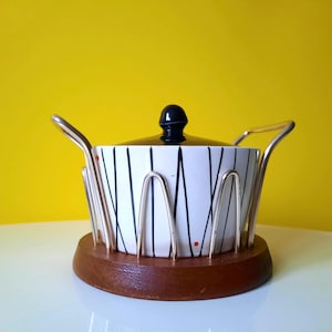 Può includere: Un piatto da portata vintage con coperchio nero e motivo a righe bianche e nere. Il piatto è posto in un supporto in metallo argentato su una base di legno. Punti rossi accentuano il design.