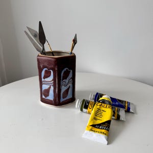 Puede incluir: Un portador de suministros de arte de cerámica granate con diseños abstractos azules y blancos, que contiene espátulas y un pincel. Tres tubos de pintura acrílica amarilla, azul y verde están sobre la mesa.