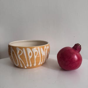 May include: A ceramic bowl with the word "DRIPPING" in white script on an orange background. A red pomegranate sits to the right of the bowl. The bowl has a white interior.