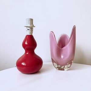 May include: A red, gourd-shaped table lamp with a white socket and a pink glass vase. The lamp has a glossy finish. The vase has a unique, organic shape and a translucent appearance. The objects are on a white surface.