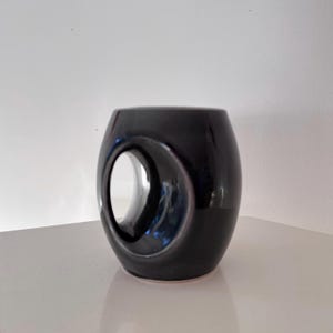 May include: A glossy black ceramic mug with a unique oval-shaped cutout. The mug has a rounded, cylindrical form and sits on a white surface. The cutout creates a distinctive design element, adding visual interest to the piece. The mug is likely used for beverages.