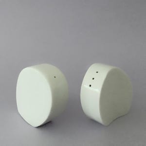 Op de afbeelding: Twee witte keramische zout- en peperstrooiers. De zoutstrooier heeft drie gaatjes, de peperstrooier één. Beide hebben een afgerond, geometrisch ontwerp tegen een grijze achtergrond.