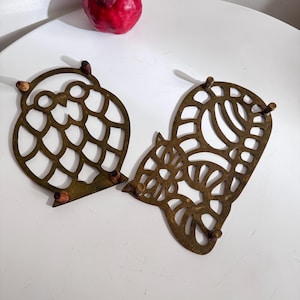 Peut inclure: Deux dessous de plat en métal couleur laiton en forme de hibou et d'écureuil. Le dessous de plat en forme de hibou a une tête circulaire et un corps avec un motif découpé. L'écureuil a un motif similaire. Les deux ont des chevilles en bois.