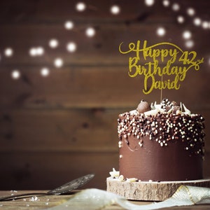 May include: A chocolate cake with chocolate sprinkles and a gold glitter cake topper that says "Happy 42nd Birthday David".