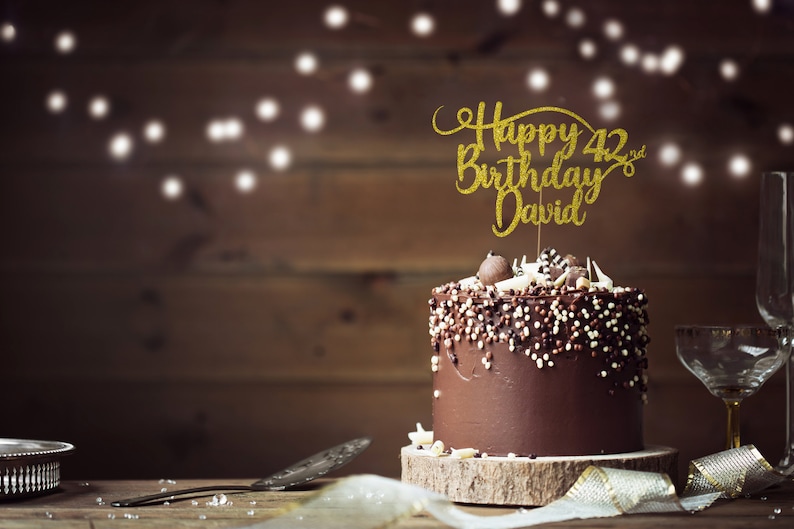 May include: A chocolate cake with chocolate sprinkles and a gold glitter cake topper that says "Happy 42nd Birthday David".