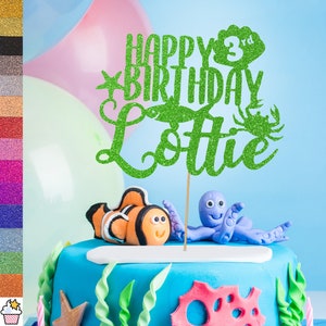 May include: Green glitter cake topper with the text "Happy 3rd Birthday Lottie" and a starfish, a crab, and a seashell.