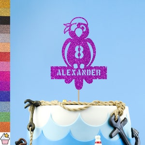May include: Purple glitter cake topper featuring a parrot design with the name "Alexander" on a banner. The parrot is wearing a bandana and has the number 8 on its chest.