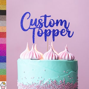 Op de afbeelding: Een blauwe glittertaarttopper met de tekst "Custom Topper" op een taart met roze glazuur en sprinkles.