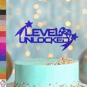 Peut inclure: Décor de gâteau en paillettes bleues avec le texte "Level Unlocked" et un graphisme de manette de jeu vidéo. Le décor est sur un gâteau glacé bleu avec des pépites colorées.