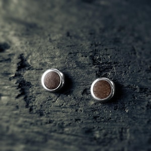 May include: A pair of silver stud earrings with round wood inlays. The wood is a dark brown color.