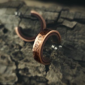 May include: A pair of copper-colored hoop earrings with a textured surface. The earrings have a dark brown interior and silver posts. The earrings are set against a blurred, textured background.