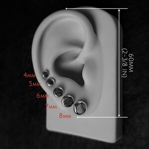 以下が含まれることがあります： 4mmから8mmのサイズ順に並べられた、ダークな質感のセンターとシルバーカラーのメタルベゼルを持つ5つの円形スタッドピアス。ディスプレイには60mmの全高が表示されています。テキストには「:BURHL ARTISAN CRAFTED ACCESSORIES」と記載されています。