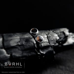 May include: A pair of circular stud earrings featuring a dark brown wood inlay set within a polished silver-toned metal frame. The text :BURHL ARTISAN CRAFTED ACCESSORIES PROUDLY HANDCRAFTED IN USA is visible in the lower left corner of the image.