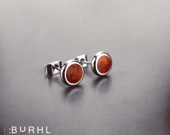 May include: A pair of stud earrings featuring circular, reddish-brown wood inlays set within polished silver-toned metal frames. The earrings have a simple, round design with a smooth, reflective finish on the metal edges and a visible wood grain.
