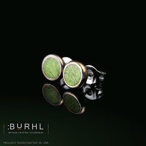 May include: A pair of stud earrings featuring circular green wood inlays set within polished gold-tone metal bezels. The earrings include metal butterfly backings and are displayed against a black background with the text :BURHL ARTISAN CRAFTED ACCESSORIES PROUDLY HANDCRAFTED IN USA.