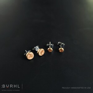 May include: Four pairs of stud earrings with a natural wood grain design. The earrings have a gold-colored bezel and silver posts. The earrings are displayed on a black background. The brand name is visible in the lower left corner.