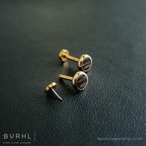 Puede incluir: Pendientes de botón dorados con un centro de veta de madera oscura. Los pendientes se muestran sobre una superficie negra texturizada. La marca ":BURHL" y el texto "ARTISAN CRAFTED ACCESSORIES" son visibles. También es visible el texto "PROUDLY HANDCRAFTED IN USA".