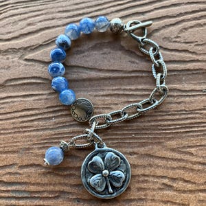 Puede incluir: Un brazalete de cadena de plata con cuentas de ágata azul, un dije de plata con un diseño de trébol de cuatro hojas y un cierre de palanca de plata.