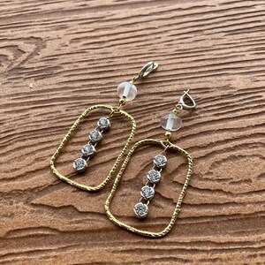 May include: A pair of gold-coloured earrings with a rectangular frame design. Each earring features a row of three clear, sparkling stones and a clear, round bead. The earrings are displayed on a wooden surface.