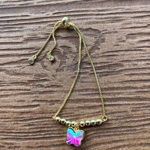 Peut inclure: Un bracelet réglable de couleur or avec un charm papillon. Le charm papillon a une finition irisée rose et bleue. Le bracelet comporte de petites perles dorées et deux petites pierres claires pendantes. Le fond est une surface en bois.