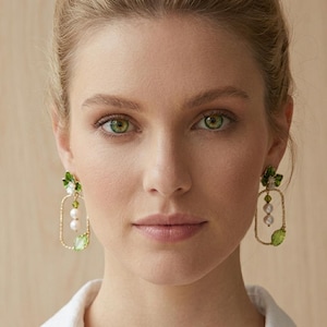 Può includere: Orecchini pendenti con una cornice rettangolare color oro, un piccolo accento a foglia verde nella parte superiore, un filo verticale centrale con due perle bianche e una piccola perlina verde, e un cristallo verde a goccia sul fondo.