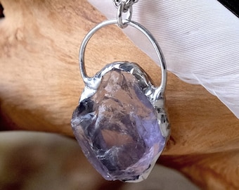Puede incluir: Un collar de cadena de plata con un colgante con un cristal de amatista en bruto. El cristal es de color púrpura claro y tiene una textura rugosa y natural. El colgante está unido a la cadena con un anillo de plata.
